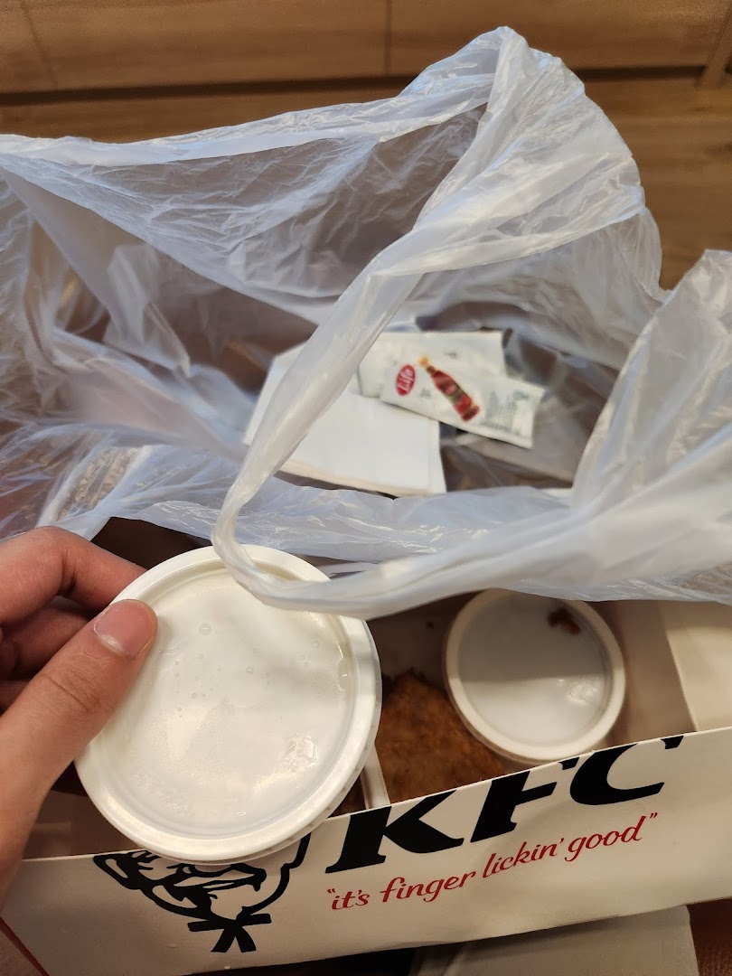 A hand holds a lid over a KFC box containing food, sauces, and a plastic bag.