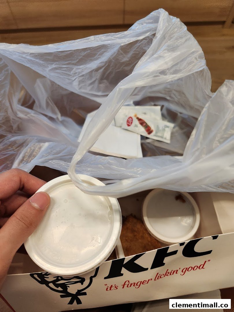 A hand holds a lid over a KFC box containing food, sauces, and a plastic bag.