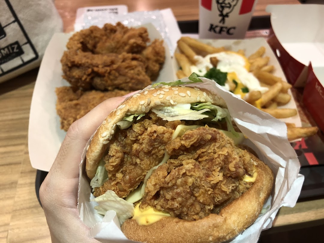 A hand holding a crispy KFC Zinger burger with fried chicken and loaded fries in the background.
