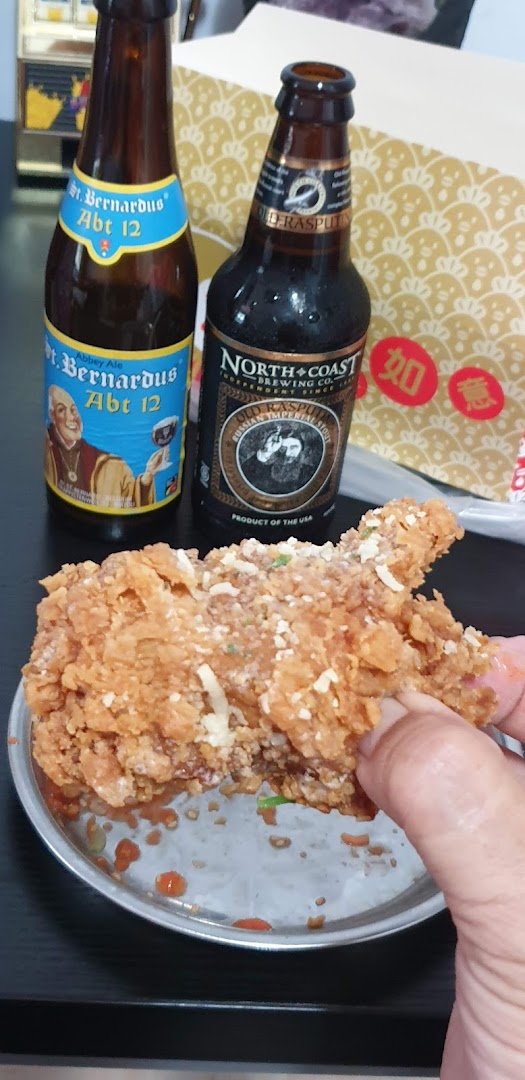 A hand holds a piece of crispy fried chicken, likely from KFC, with two beer bottles in the background.