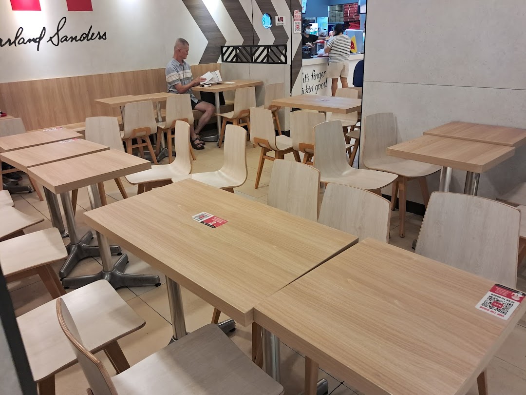 The image shows the clean and modern dining area of KFC, with light wood tables and chairs, and a customer seated.