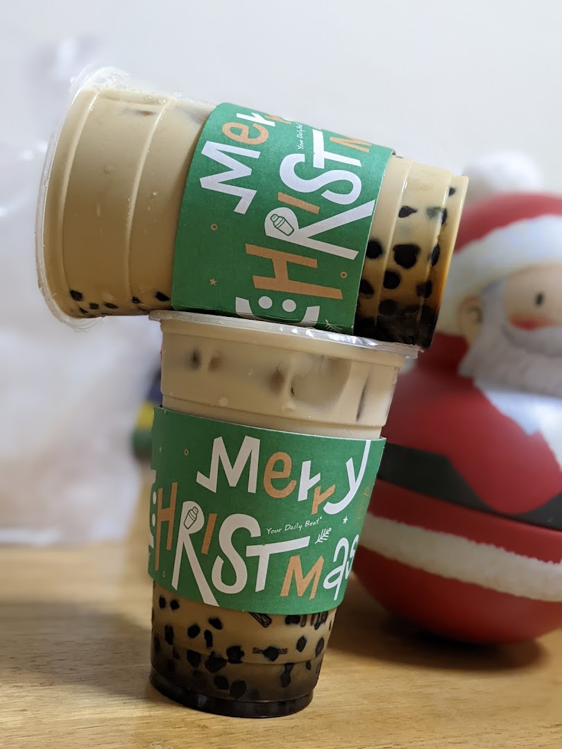 Two bubble tea drinks with festive Christmas sleeves are stacked, with a Santa figurine nearby.