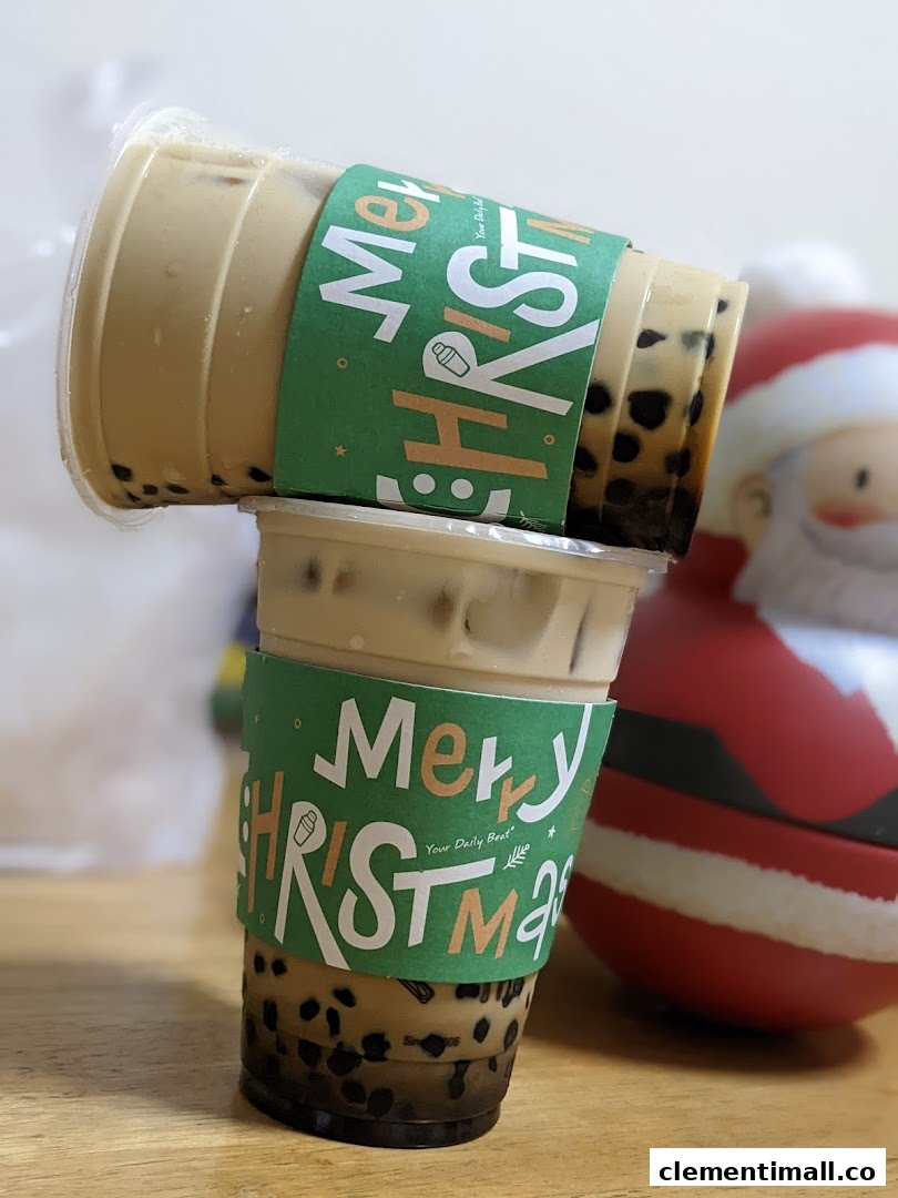 Two bubble tea drinks with festive Christmas sleeves are stacked, with a Santa figurine nearby.