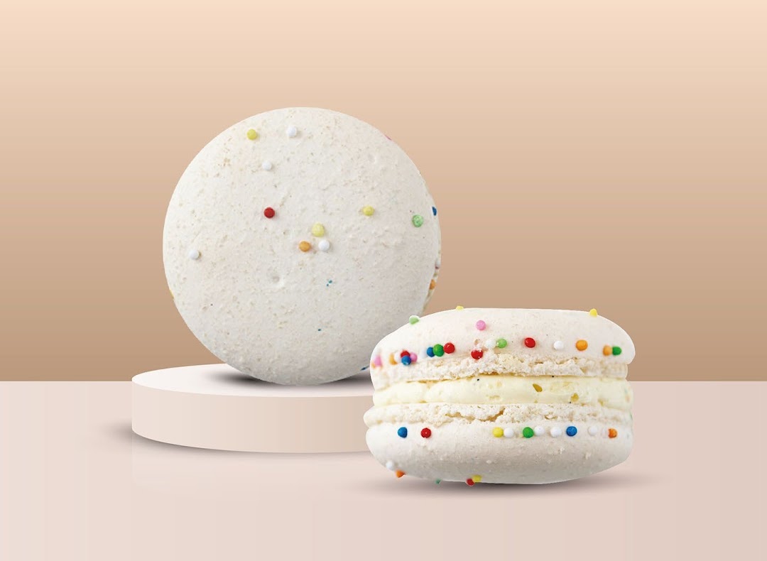 Two white macarons with colorful sprinkles, one on a pedestal, against a light background.