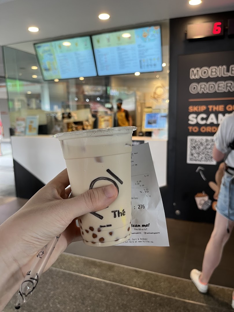 A hand holds a KOI Thé bubble tea drink with the shop counter and menu screens in the background.