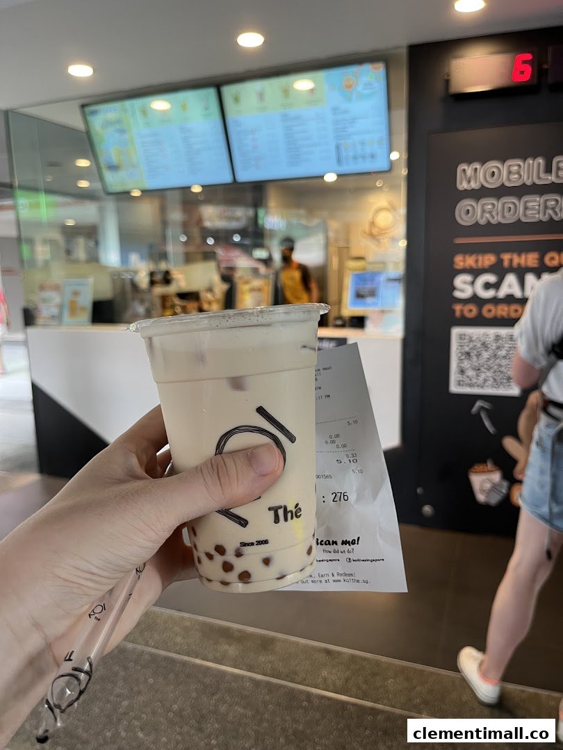 A hand holds a KOI Thé bubble tea drink with the shop counter and menu screens in the background.