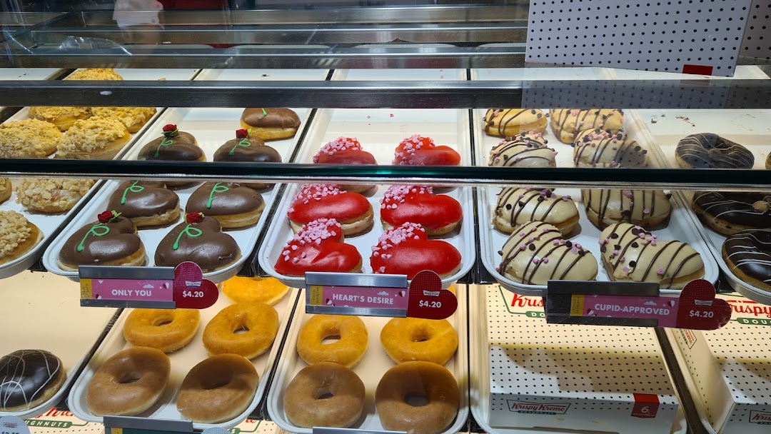 A vibrant display of Krispy Kreme donuts, including special heart-shaped varieties, in a glass case.