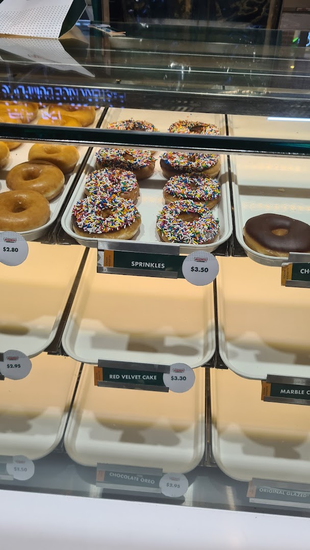 A Krispy Kreme display case showcasing various donuts like Original Glazed, Sprinkles, Red Velvet Cake, and Chocolate Oreo with prices.
