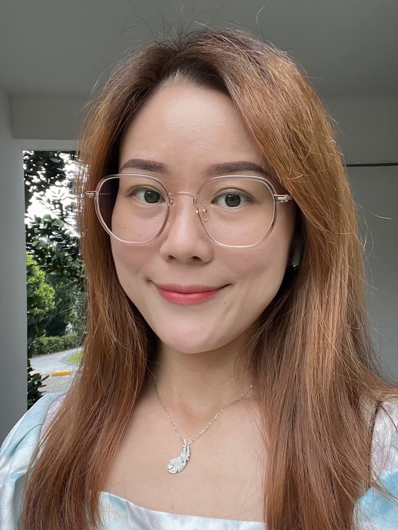 A close-up portrait of a smiling woman wearing glasses and a feather-shaped necklace.