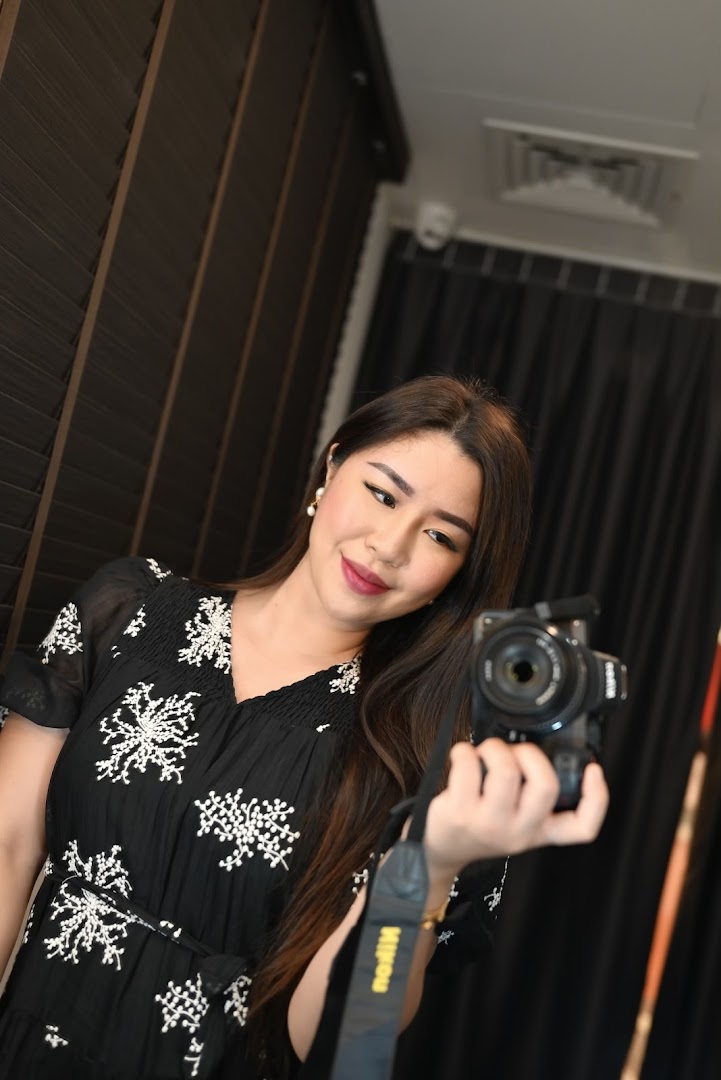 A woman in a black dress with white embroidery holds a camera, possibly taking a selfie.