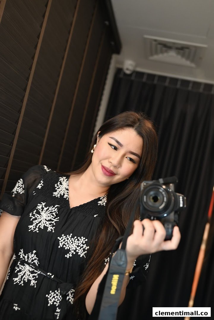 A woman in a black dress with white embroidery holds a camera, possibly taking a selfie.