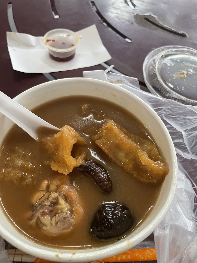 A close-up of a bowl of herbal soup with chicken, fried dough, and dark ingredients on a table.