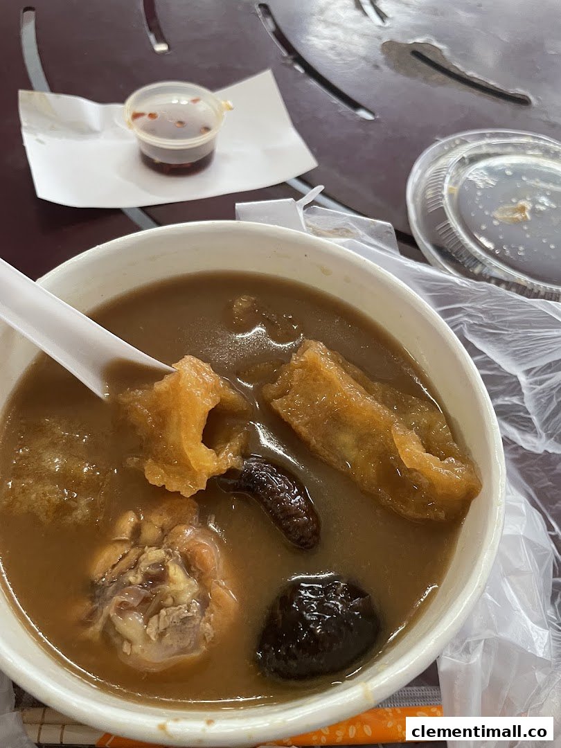A close-up of a bowl of herbal soup with chicken, fried dough, and dark ingredients on a table.