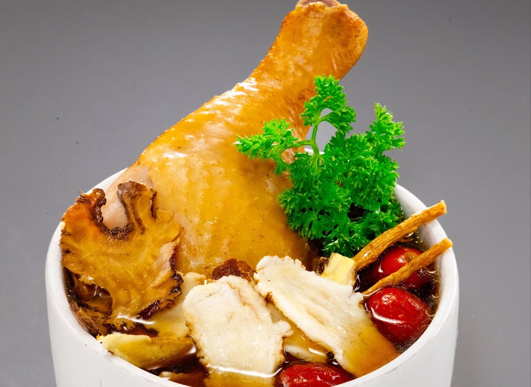 A close-up of a bowl of herbal soup featuring a chicken drumstick, red dates, and various traditional herbs.