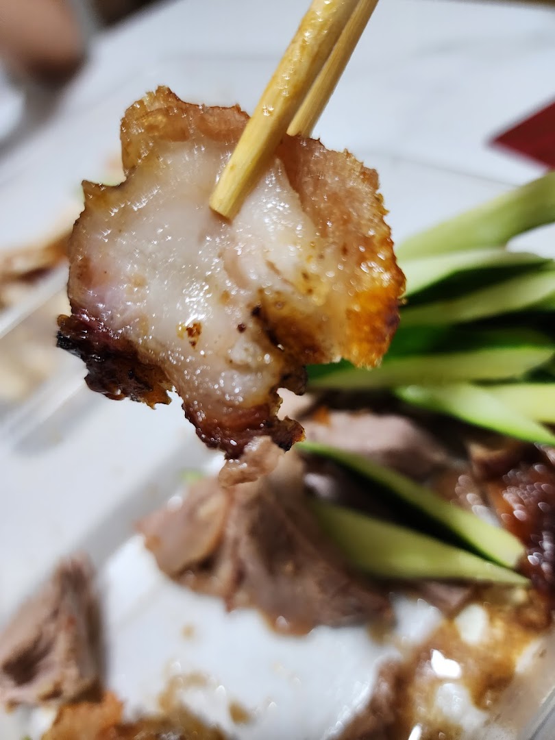 A close-up of a crispy piece of duck held by chopsticks, with more duck and cucumber in the background.