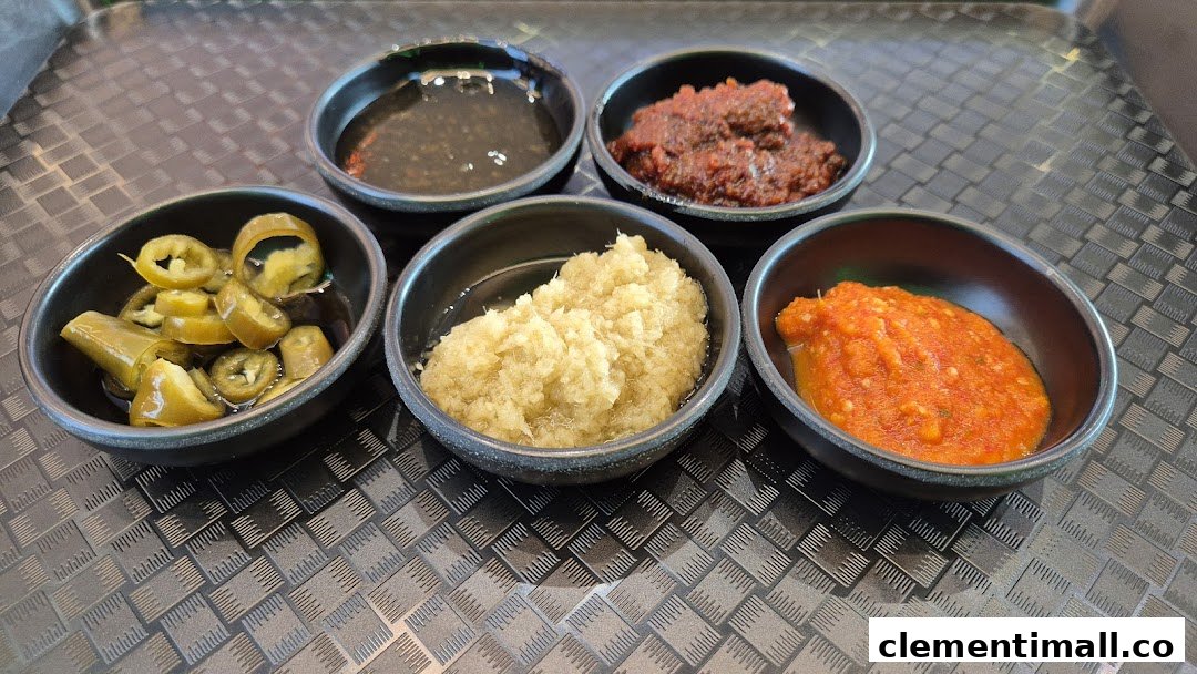 Five small bowls of various flavorful condiments including chili, garlic, and sauces on a textured tray.