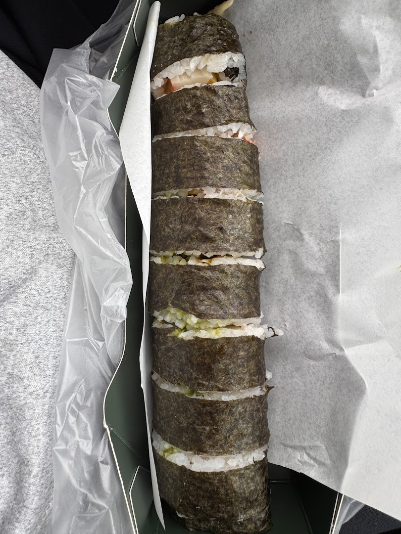 A long, cut sushi roll with various fillings, presented in a takeaway box from Maki-San.