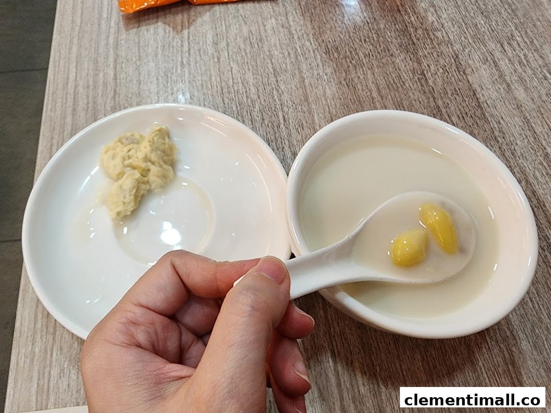 A hand holds a spoon with ginkgo nuts in a sweet soup, alongside a plate of mashed taro dessert.