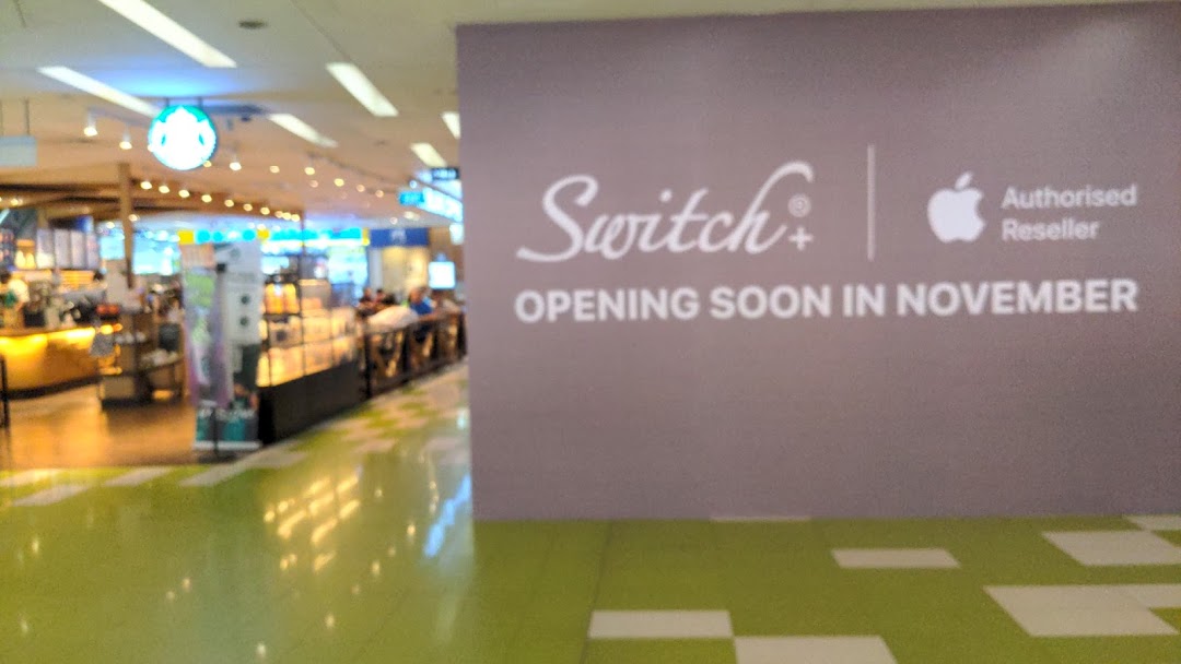 A mall scene featuring a 'Switch+' Apple Reseller 'Opening Soon' sign, with a Starbucks store in the background.