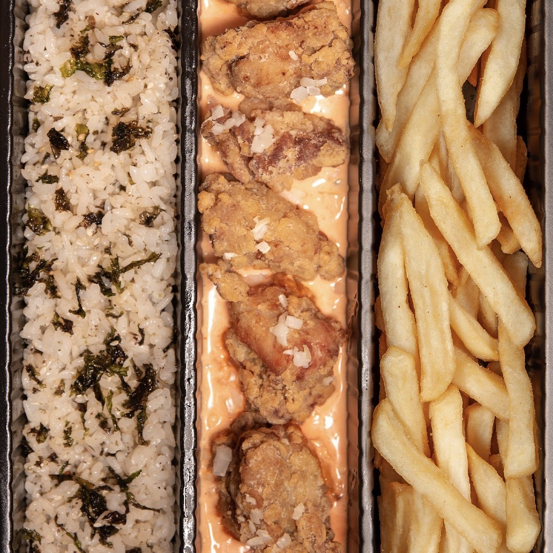 A close-up of a bento box featuring fried chicken, seaweed rice, and golden fries.