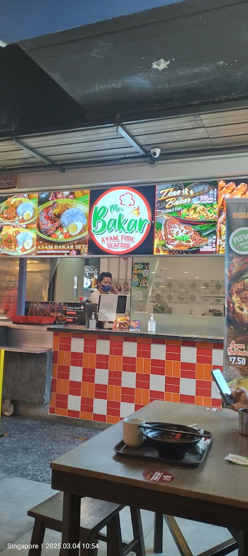 A staff member behind the counter at Mr Bakar, with vibrant menu displays and a customer table.