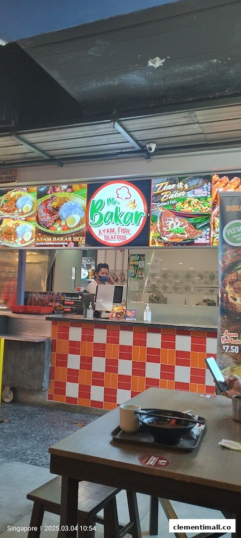 A staff member behind the counter at Mr Bakar, with vibrant menu displays and a customer table.
