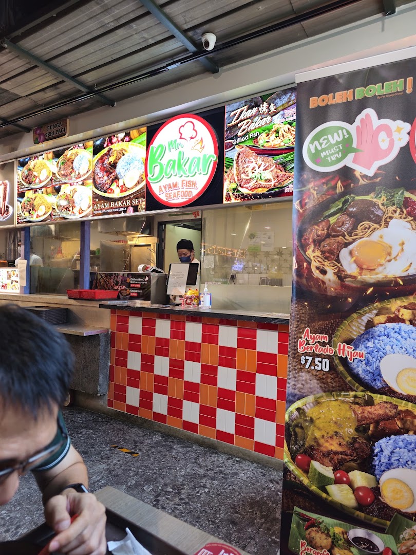 View of Mr Bakar food stall counter featuring food display boards, logo, and a staff member.