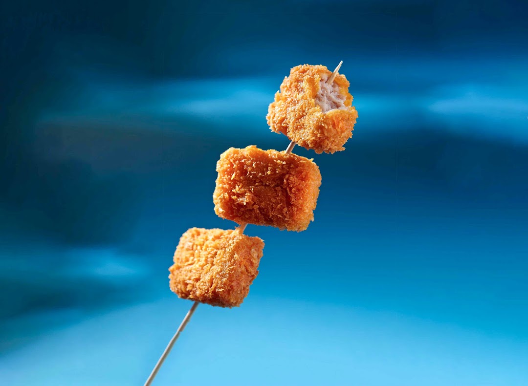 Three golden-fried nuggets on a skewer against a vibrant blue background, with one bitten.