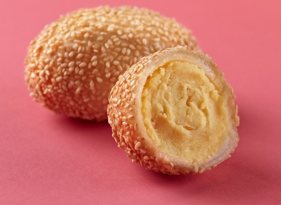 A golden sesame ball, one cut open to reveal its sweet, yellow filling, on a pink background.
