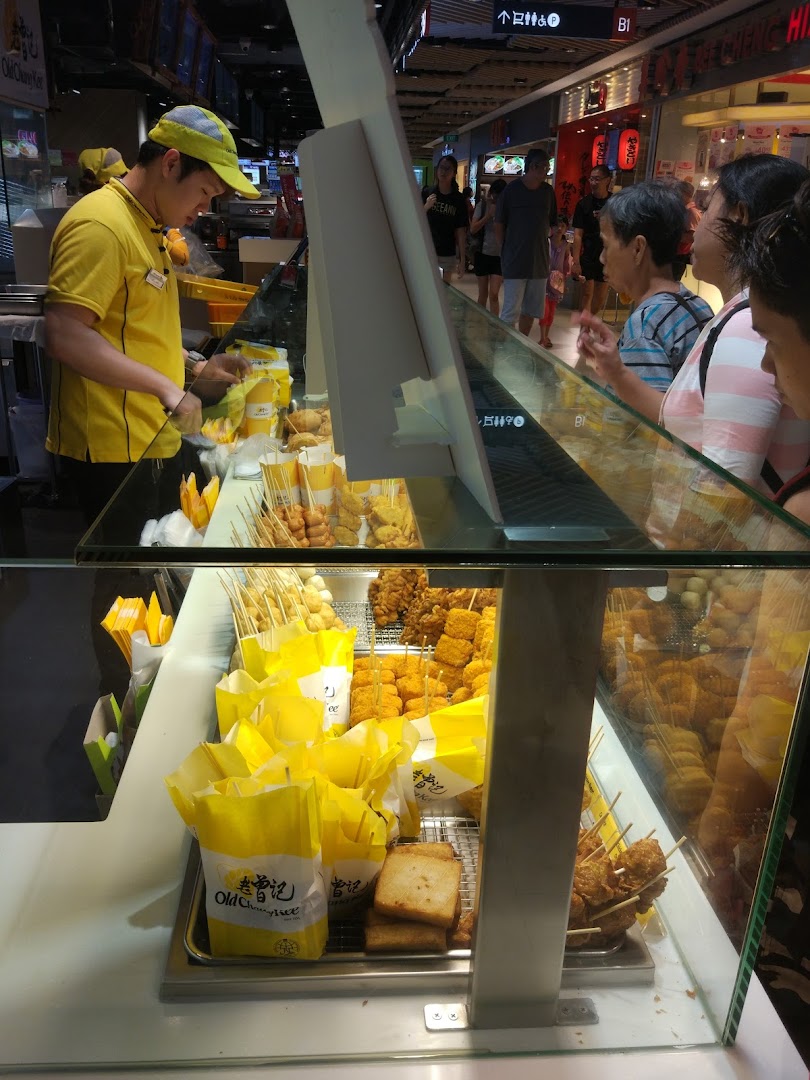 A staff member serves customers at Old Chang Kee, showcasing a variety of freshly fried snacks in a display counter.
