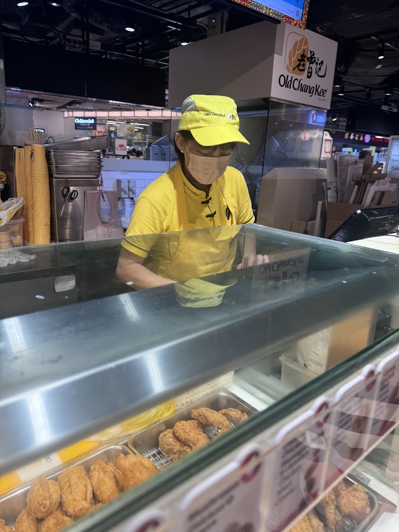 An Old Chang Kee staff member behind a counter with various fried snacks on display.
