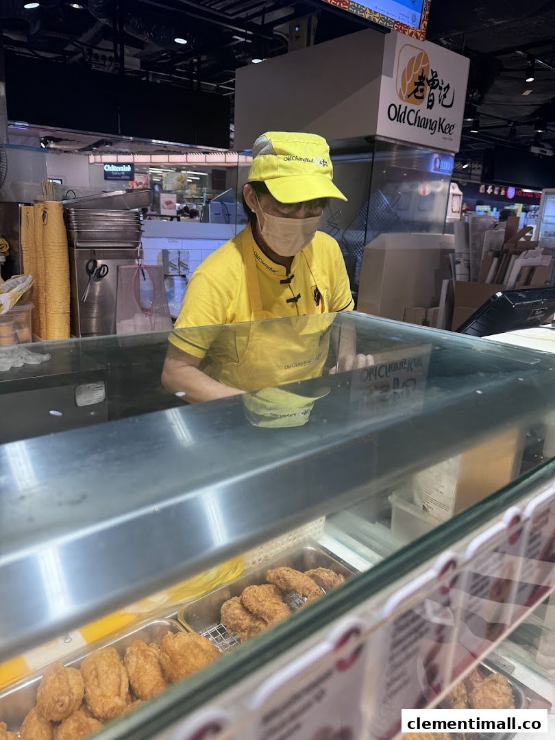An Old Chang Kee staff member behind a counter with various fried snacks on display.