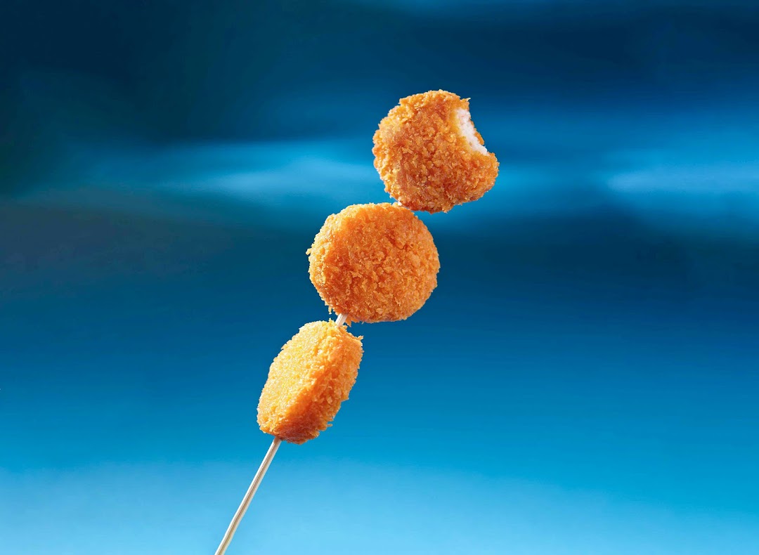 Three golden-fried items on a skewer against a vibrant blue background, one with a bite taken.