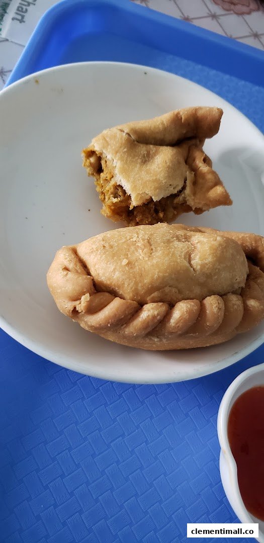 Two golden-brown curry puffs on a white plate, one revealing its savory filling, with a side of chili sauce.