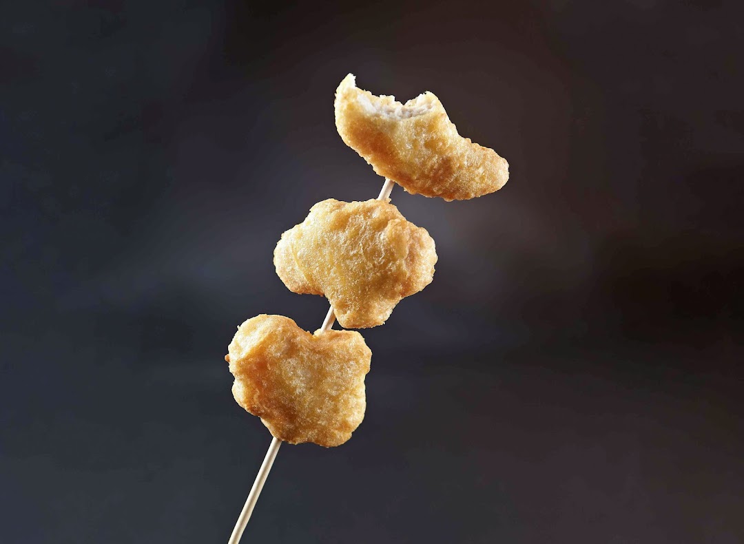 Three golden-brown fried snacks, possibly chicken nuggets, on a skewer against a dark background.