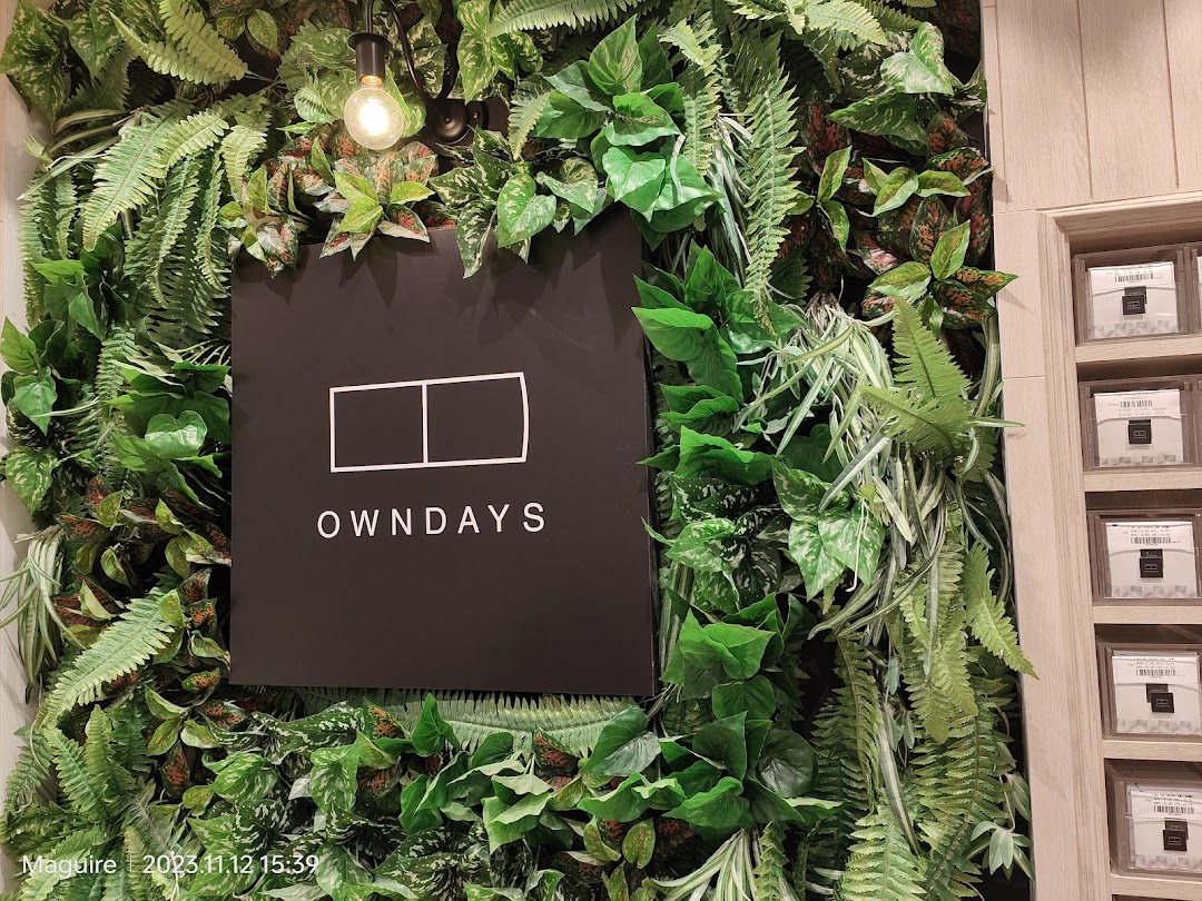 Close-up of the OWNDAYS logo sign set against a lush green artificial plant wall.