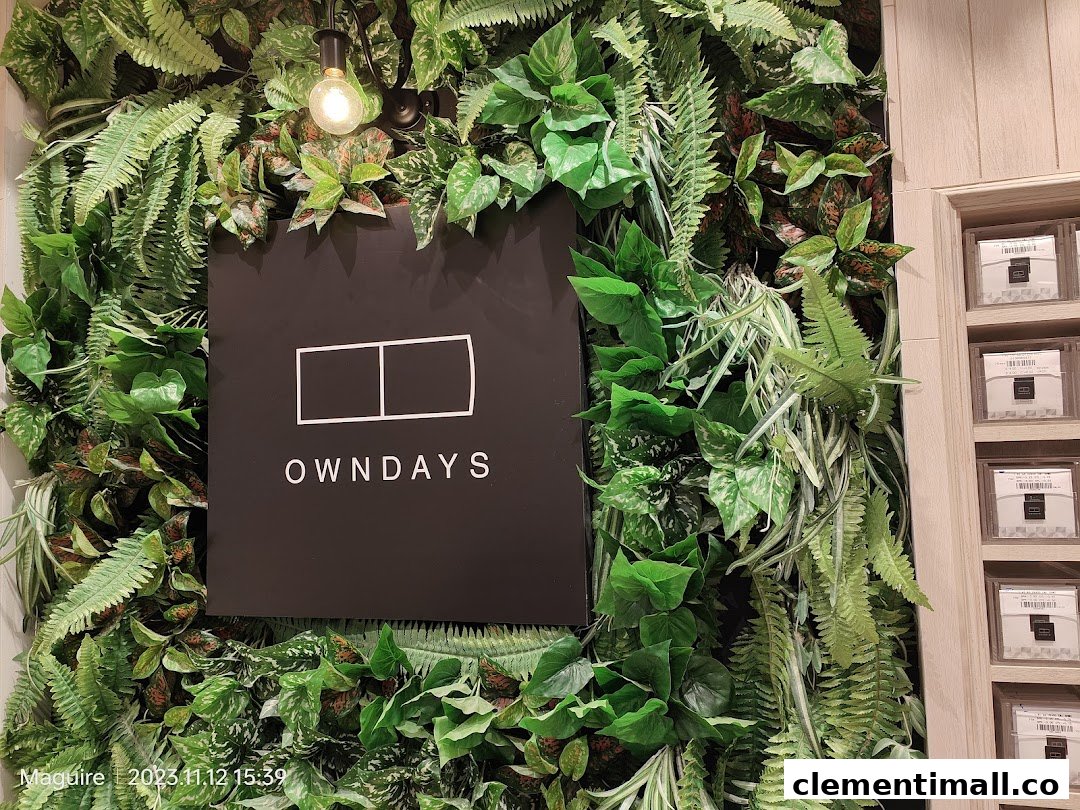 Close-up of the OWNDAYS logo sign set against a lush green artificial plant wall.