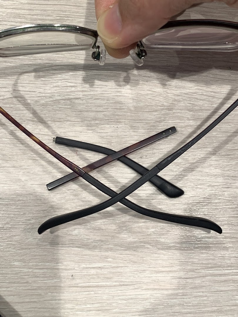 A hand holds eyeglasses, with detached temples lying on a wooden surface, suggesting repair or assembly.