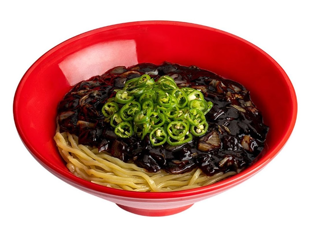 A vibrant red bowl filled with Jajangmyeon noodles, topped with rich black bean sauce and fresh green chili slices.