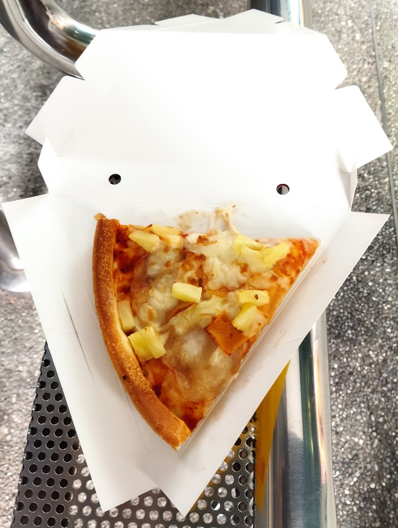 A delicious slice of Hawaiian pizza with pineapple and ham in a takeaway box.