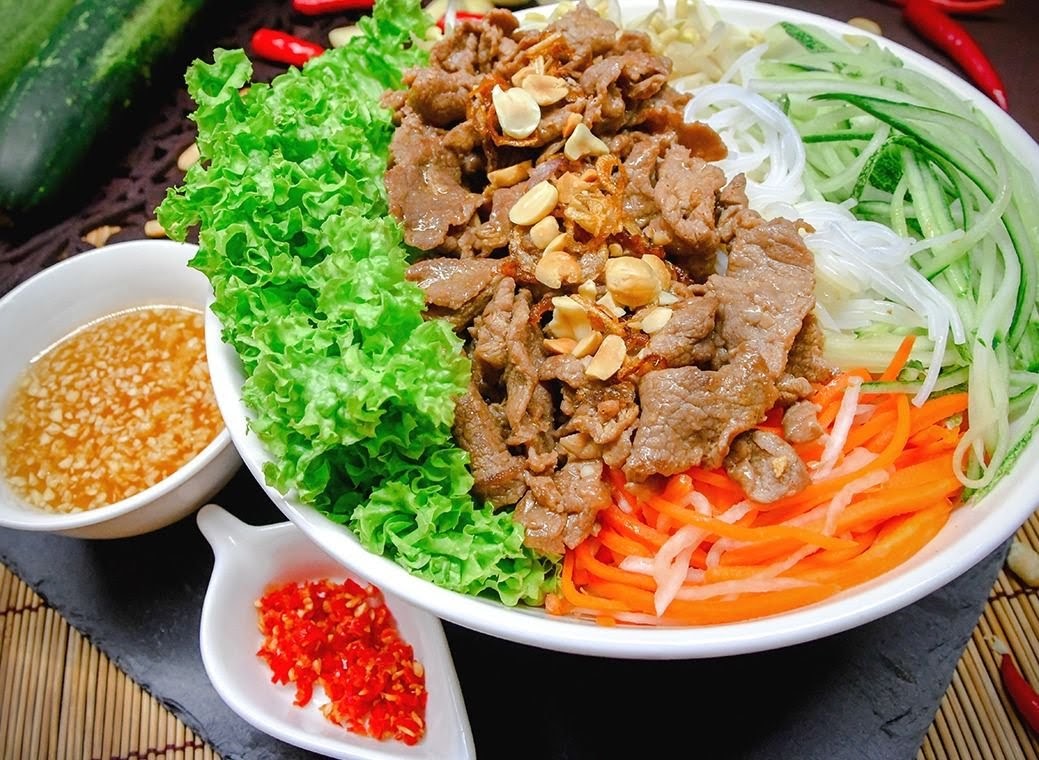 A vibrant bowl of Vietnamese beef noodle salad with fresh vegetables and a side of sauce.