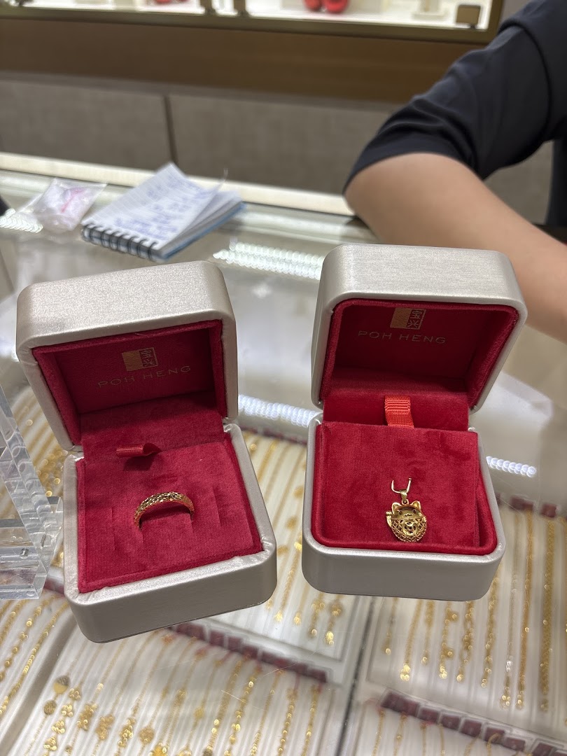 Two open Poh Heng jewellery boxes displaying a gold ring and a gold pendant on a counter.