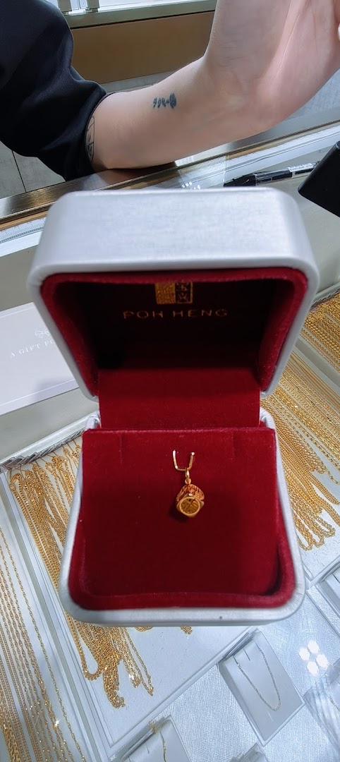 A delicate gold charm displayed in a red velvet Poh Heng jewelry box, surrounded by gold chains.