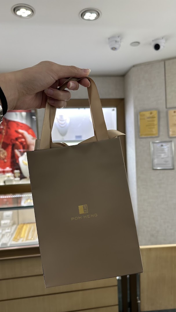 A hand holds a Poh Heng Jewellery shopping bag inside the store, with display cases visible in the background.