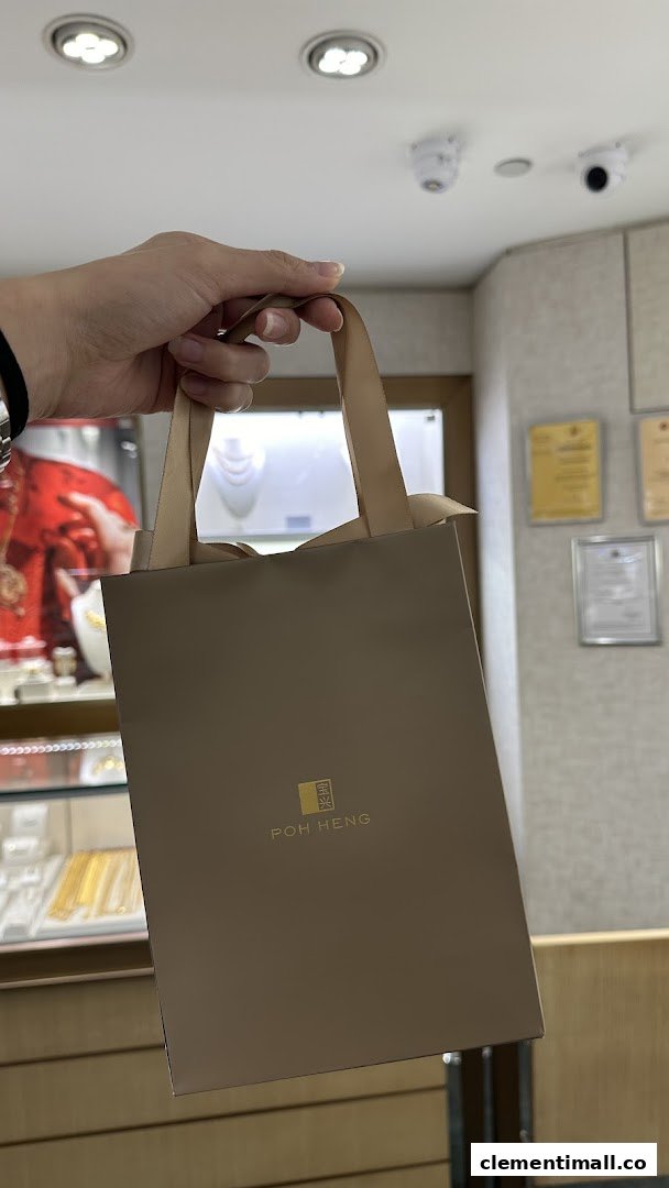 A hand holds a Poh Heng Jewellery shopping bag inside the store, with display cases visible in the background.
