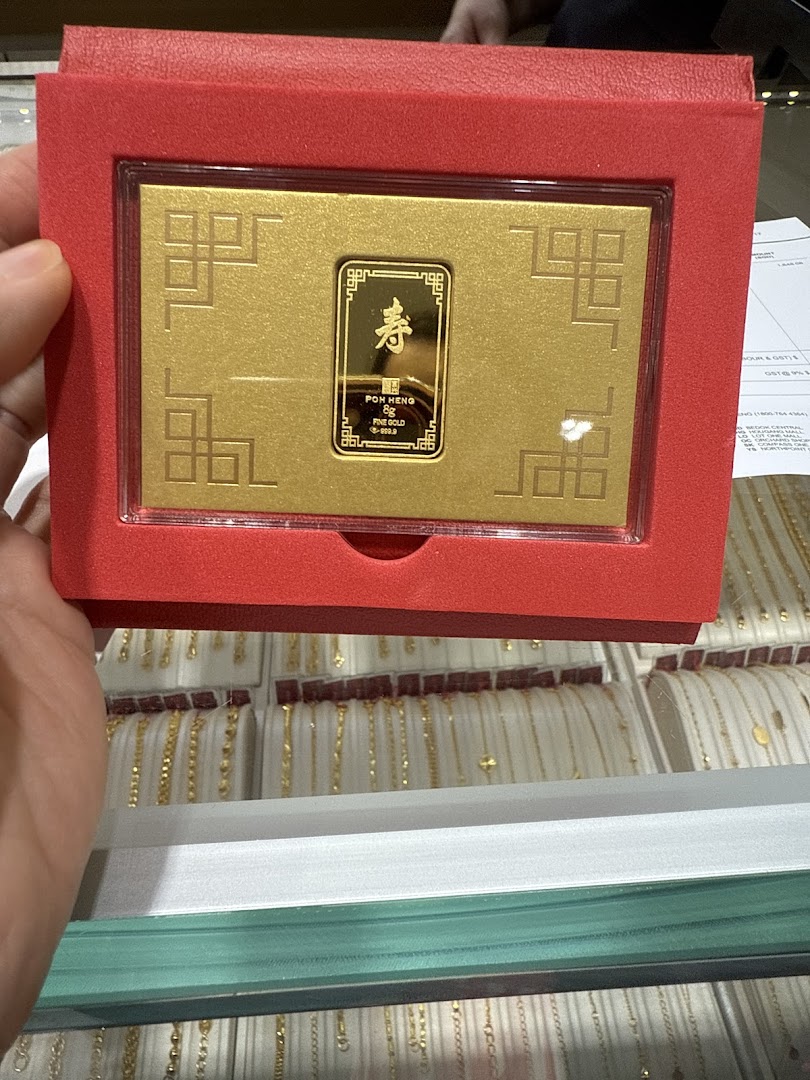 A hand holds a red box showcasing a Poh Heng 8g fine gold bar, with other gold jewelry in display cases.