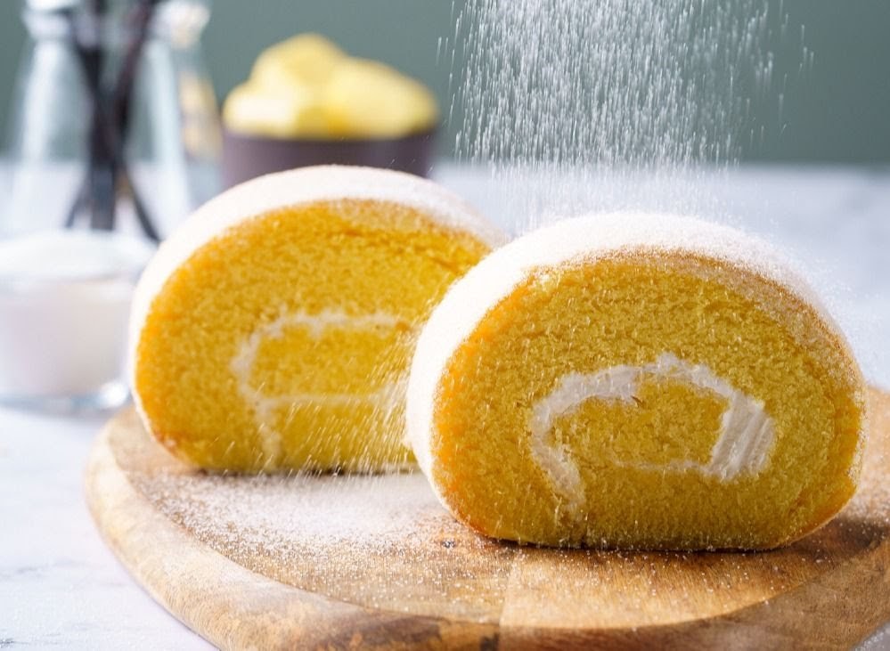 Two golden cake rolls being dusted with powdered sugar on a wooden board.