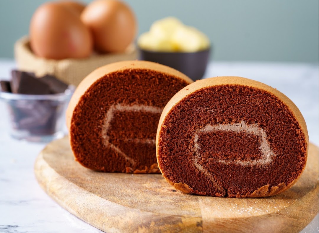 Two slices of chocolate cake roll on a wooden board, with baking ingredients in the background.