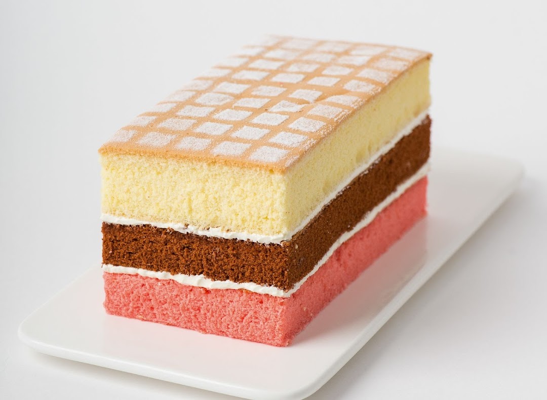 A rectangular layered cake with vanilla, chocolate, and strawberry sponges on a white plate.