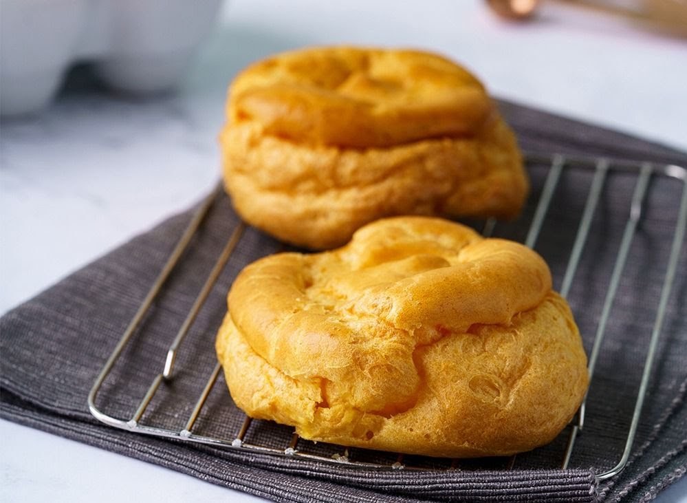 Two golden-brown cream puffs resting on a cooling rack with a dark grey cloth.